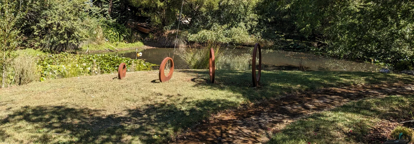 Rusted steel ring sculptures on a lawn beside a garden pond, an old rowboat moored at the bank