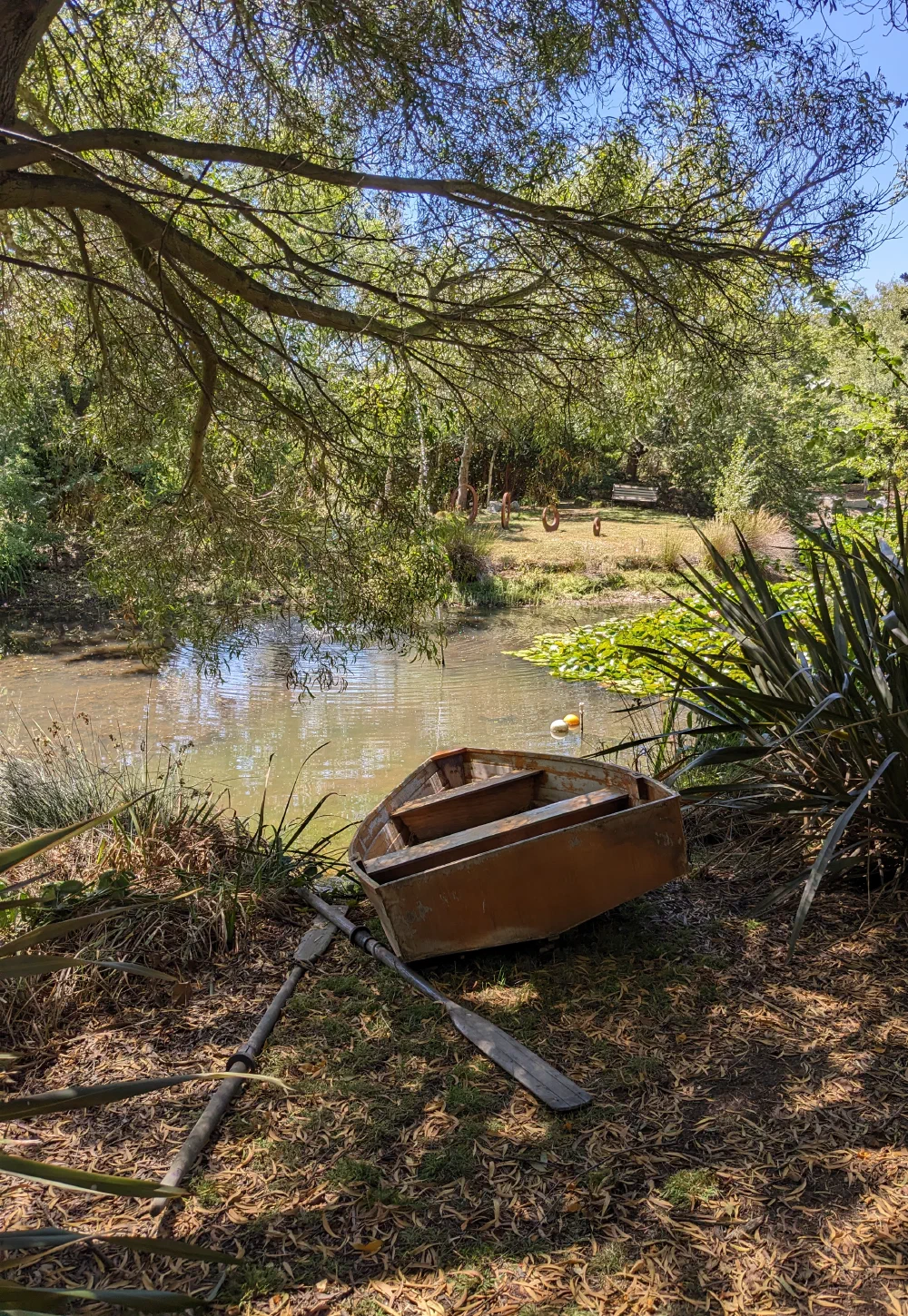 A weathered wooden rowboat and oars pulled up on the bank, ring sculptures across the water
