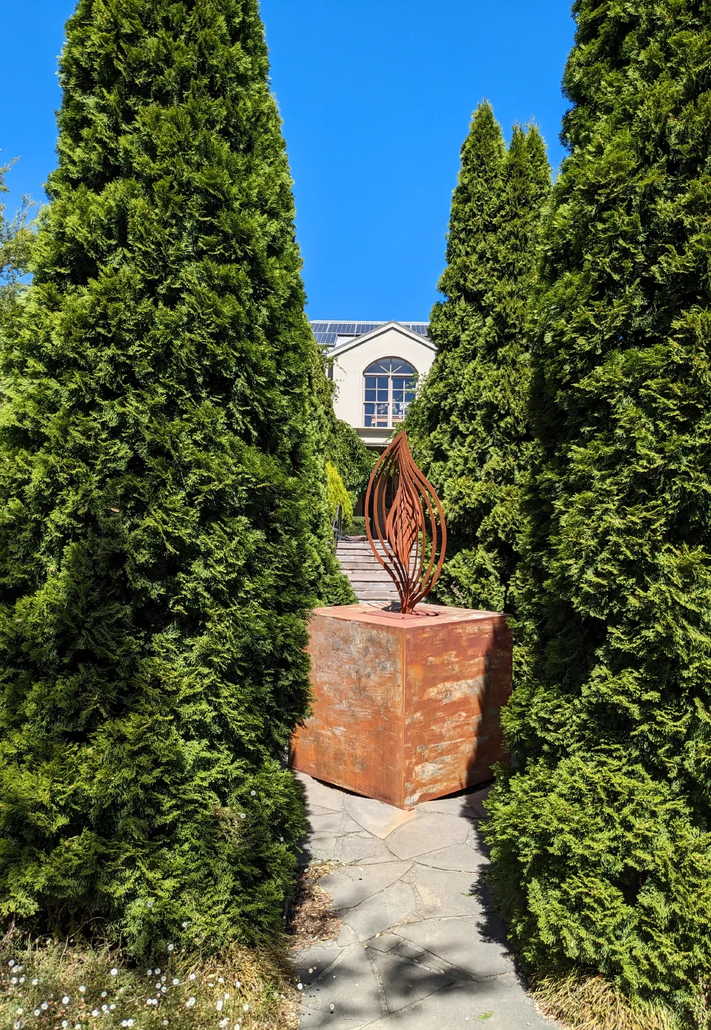 A flame-shaped corten steel sculpture on a rusted plinth, framed by a clipped conifer hedge