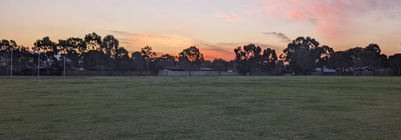 Sunset colours streaking pink and orange above a football oval's treeline