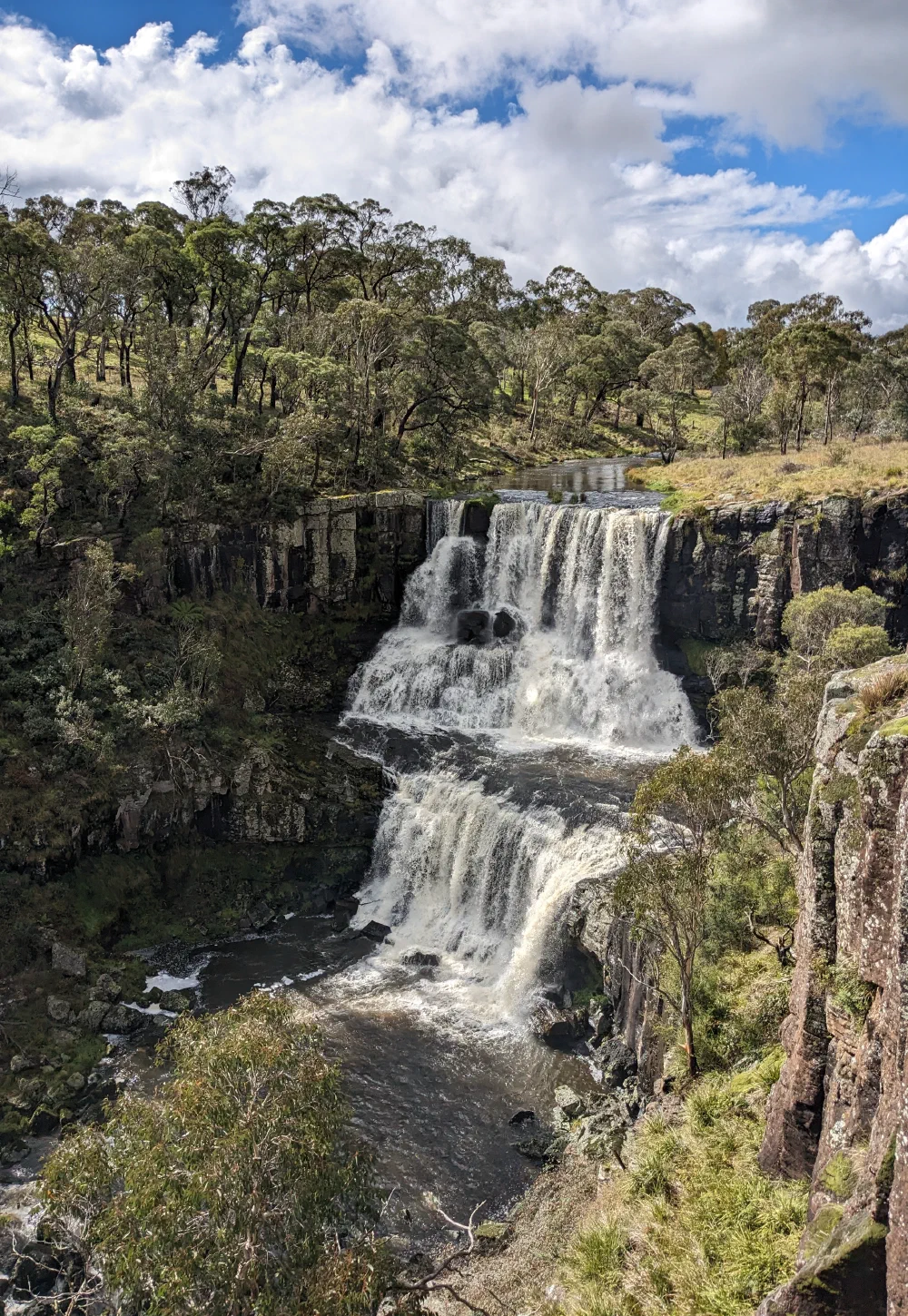 A two-tiered waterfall thundering over a basalt cliff into a forested gorge